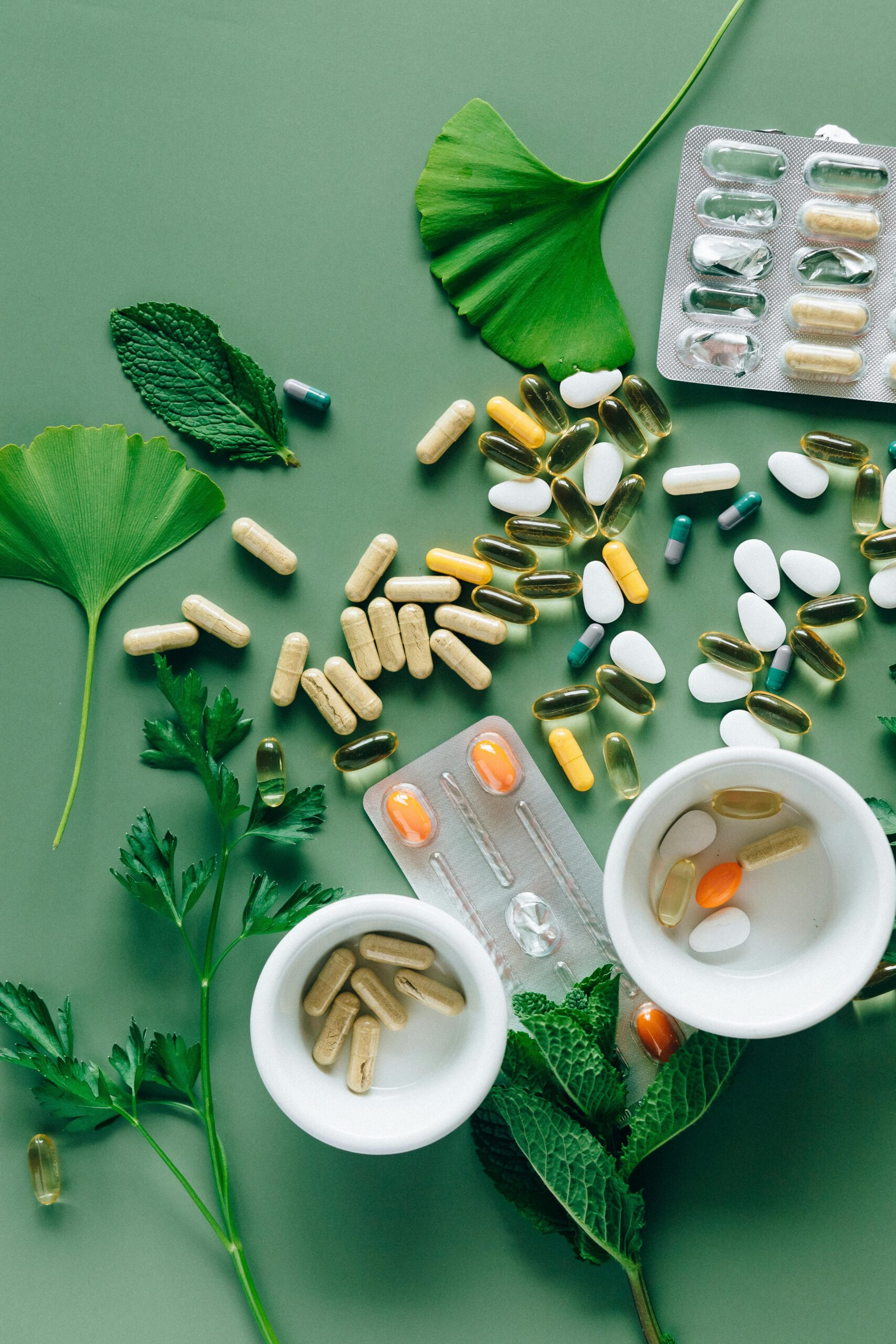 Flat lay of assorted pills and herbs on green surface, symbolizing health and wellness.