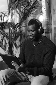 Black and white portrait of a man wearing headphones and using a tablet in a cozy indoor setting.